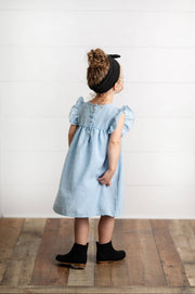 Child wearing a light blue dress with ruffled sleeves and black boots on a wooden floor.