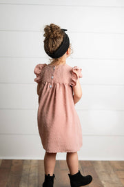 Child wearing a pink dress with ruffled sleeves against a white wooden panel background