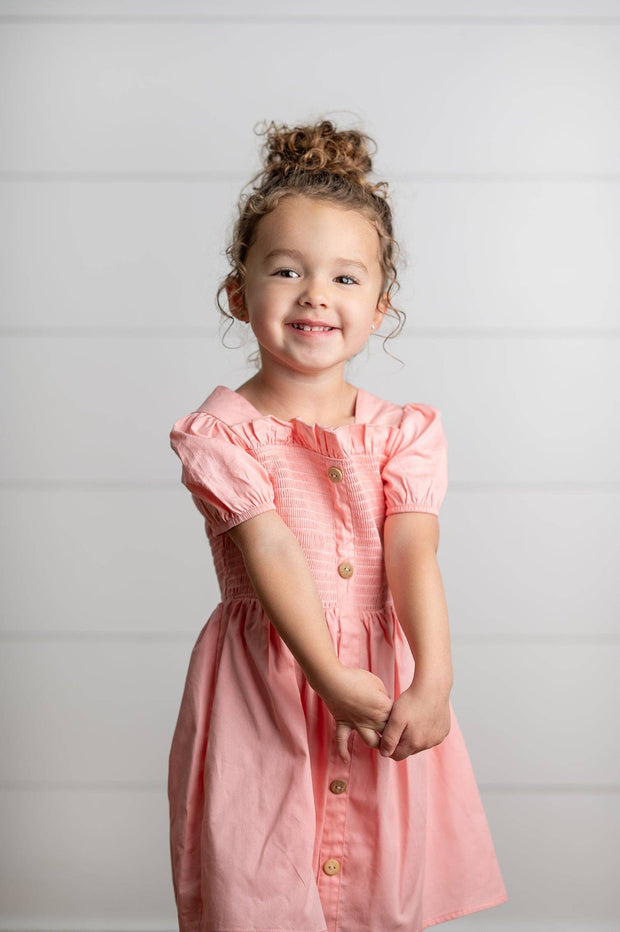 Young girl wearing a pink dress with buttons on a plain background