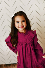 Young girl wearing a burgundy dress with ruffled details against a white geometric-patterned wall.