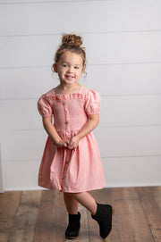 Young girl wearing a pink dress standing against a white wall.