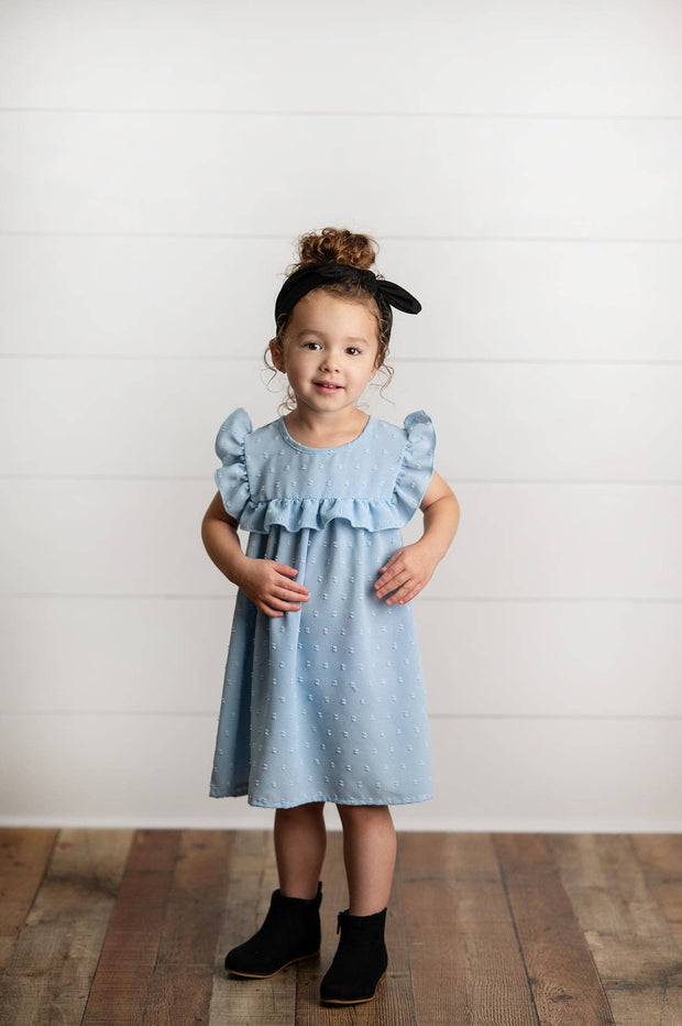 Young girl wearing a light blue dress with ruffled sleeves against a white wooden panel background.