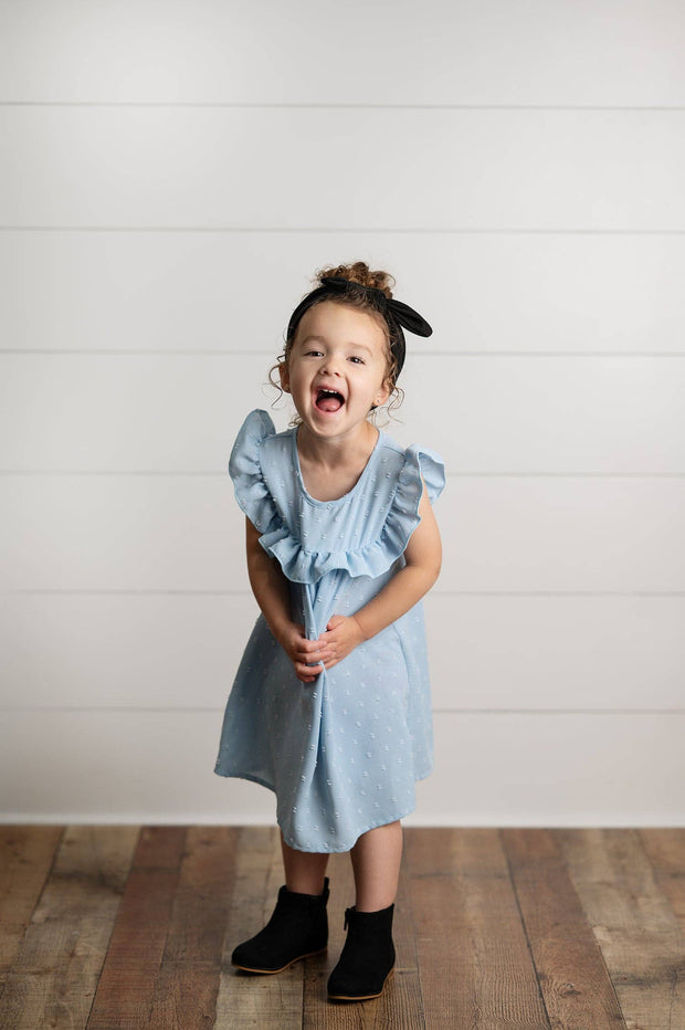 Child wearing a light blue dress with ruffled sleeves against a white wooden panel background
