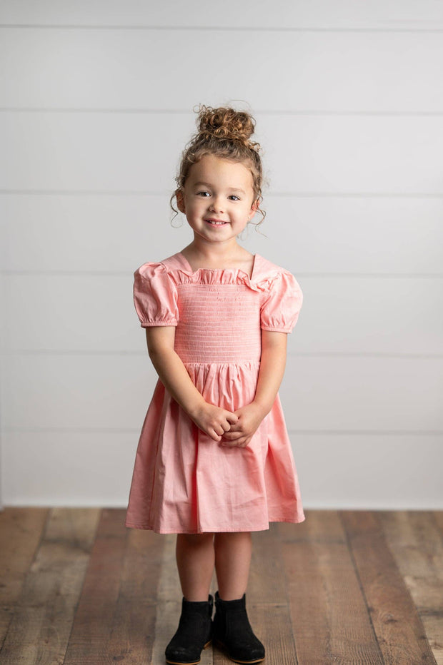 Young girl wearing a pink dress standing on a wooden floor with a white wall background
