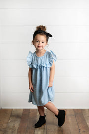 Young girl wearing a light blue dress with ruffled sleeves against a white wall.