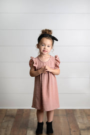 Young girl wearing a pink dress standing against a white wooden panel background